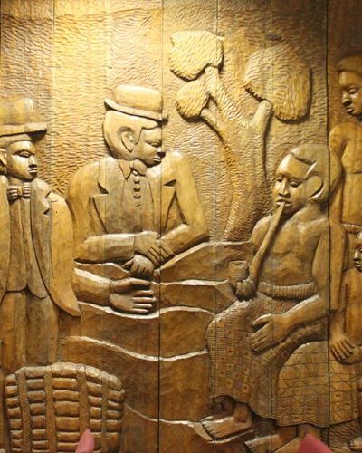 A carved wooden panel depicts a detailed scene of five people. Two men in suits and hats are shaking hands. The other three figures, including a seated woman holding a pipe and two children, observe. A tree is carved in the background.