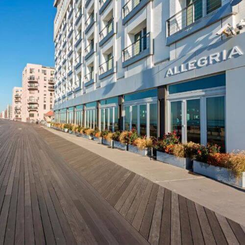 A modern beachfront hotel with large windows and a wooden boardwalk lined with plants. The clear blue sky reflects the sunny weather. A row of buildings extends into the distance along the boardwalk.