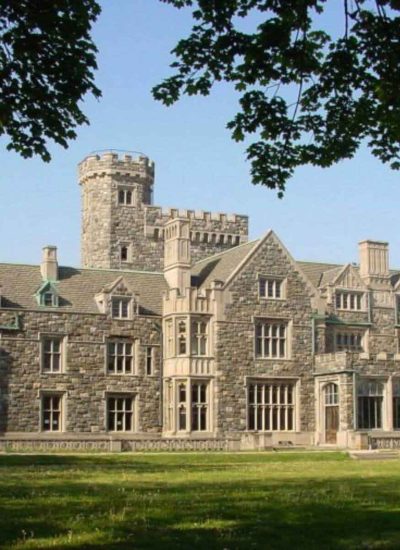 A grand stone mansion in Sands Point boasts multiple chimneys and a central castle-like tower, surrounded by trees and lush greenery under a clear blue sky. The architecture features large windows and intricate stonework details.