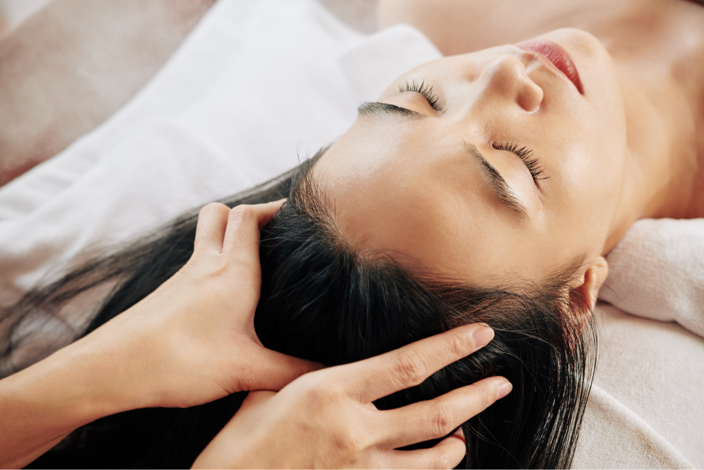 A woman lies with her eyes closed, receiving a relaxing head massage. Her dark hair is spread out on a white towel, and the hands of the masseuse gently cradle her head. The scene conveys tranquility and relaxation.
