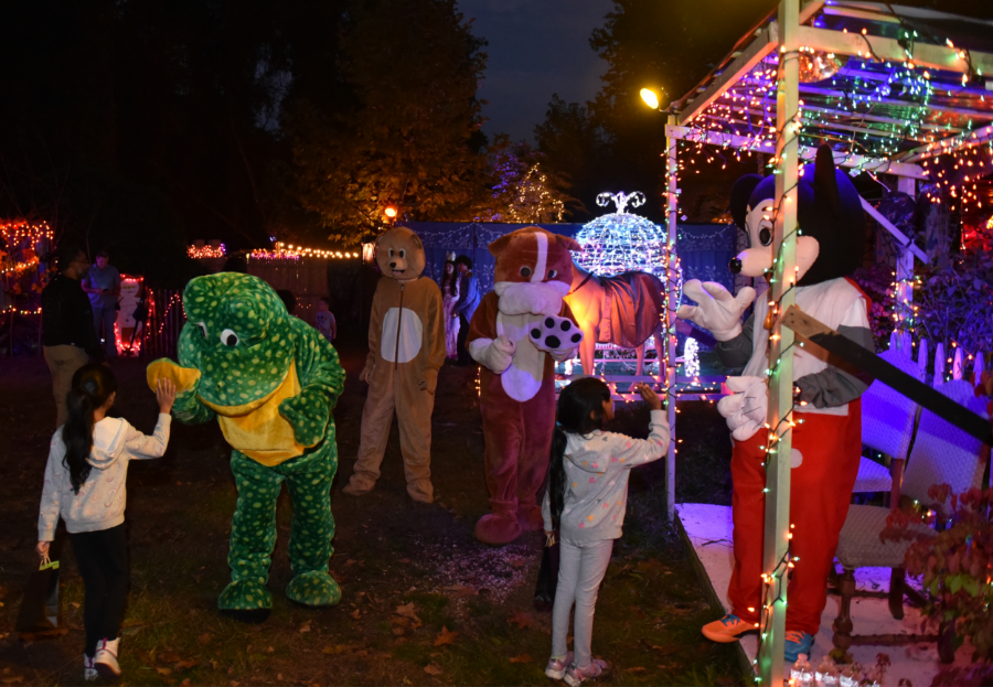 Children interact with people in colorful animal costumes, including a green dinosaur and a brown dog, under festive lights at night. A costumed character resembling a well-known mouse stands nearby, creating a joyful, lively atmosphere.