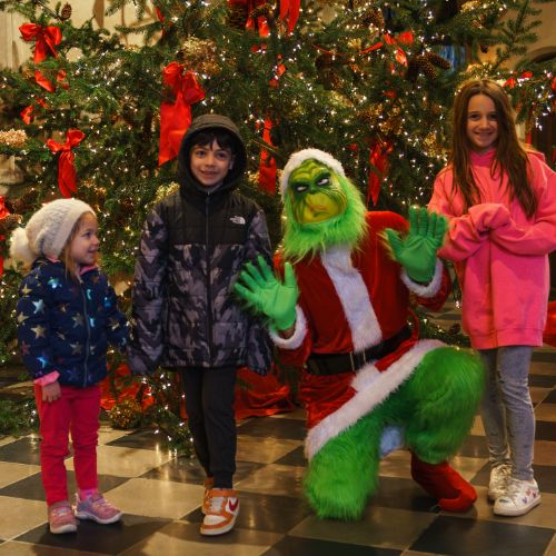 Three children standing next to a person dressed as the Grinch in front of a decorated Christmas tree. The kids are smiling, and the Grinch is posing playfully with exaggerated gestures. The scene is festive and colorful.