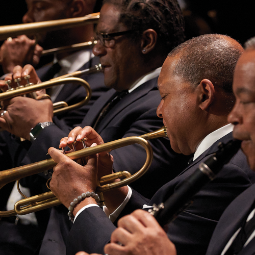 A group of musicians in suits perform, focusing on trumpet and clarinet players. The brass instruments shine under stage lights, and the musicians are deeply engaged in their performance.