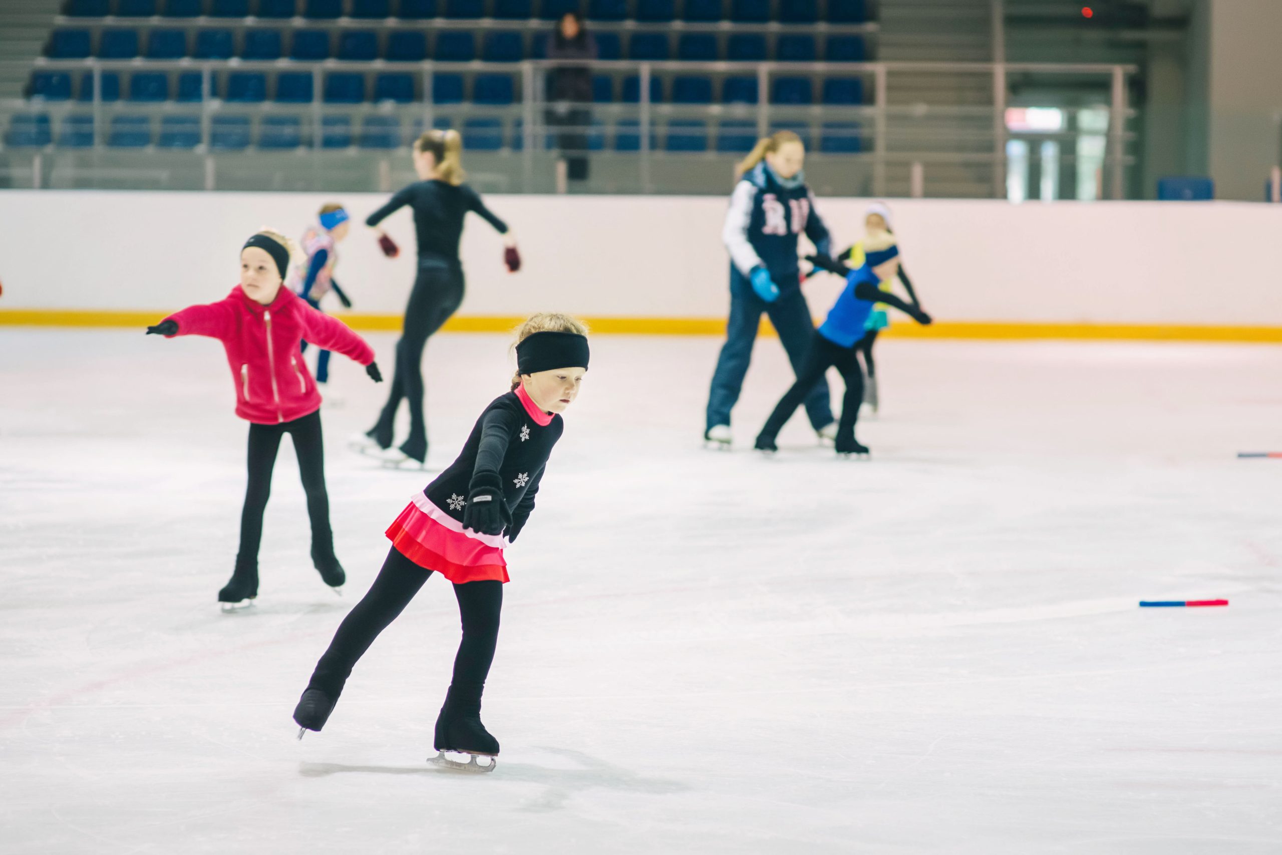Children practice figure skating on an indoor ice rink. A young girl in a black and red outfit is in the foreground, while other skaters and a coach are seen in the background on the ice. The rink has empty seating.