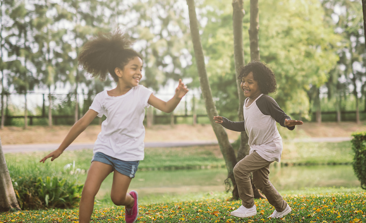 Two children joyfully running and playing in a park on a sunny day. They are surrounded by green grass and trees, with a small pond in the background. Both are smiling and appear carefree.