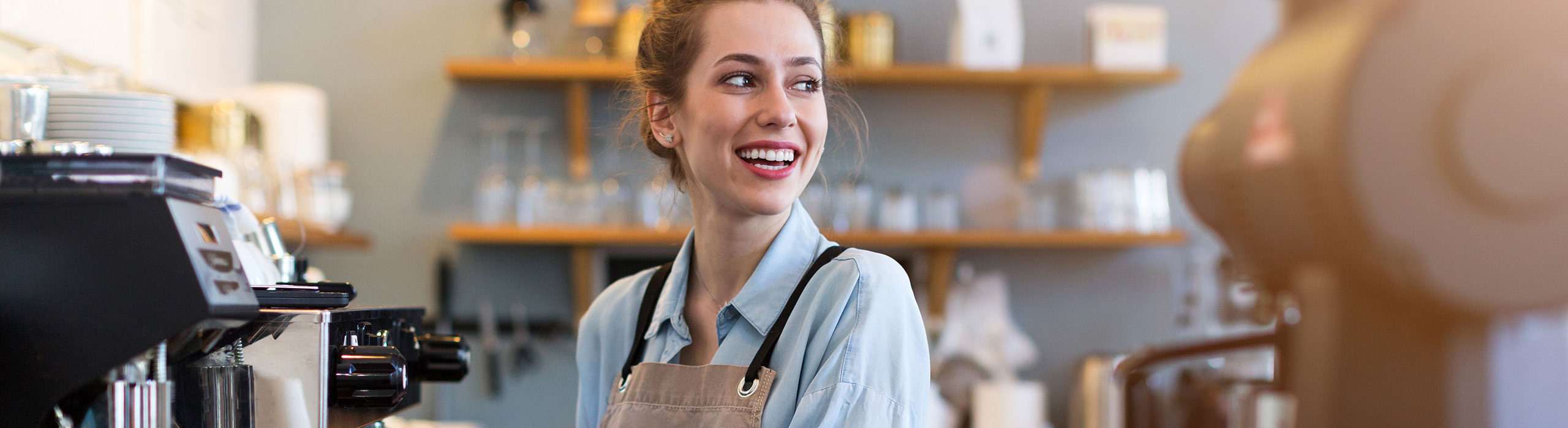 A smiling barista wearing a beige apron stands behind a coffee machine in a cozy café. Shelves with cups and jars are visible in the background.