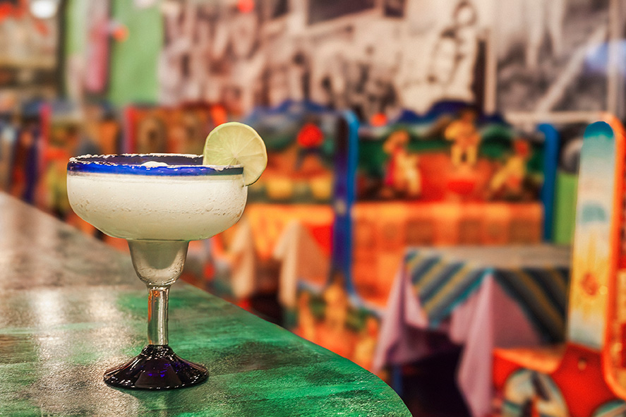 A margarita with a salted rim and lime wedge sits on a green countertop in a colorful, festive restaurant with vibrant, painted tables and chairs in the background.