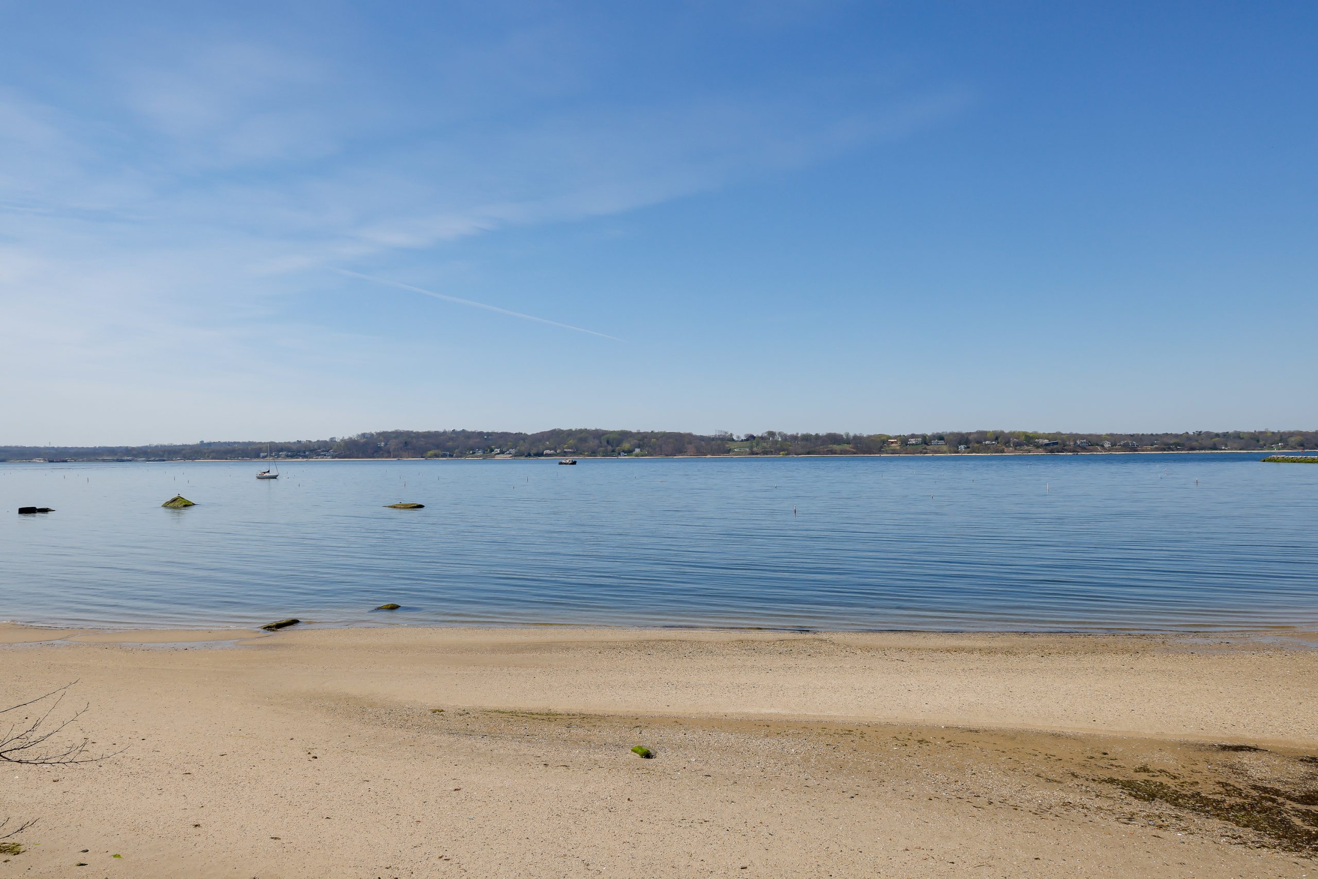 A sandy beach with calm blue water under a clear sky. A few small boats float on the water, and the distant shoreline is lined with trees and low hills.