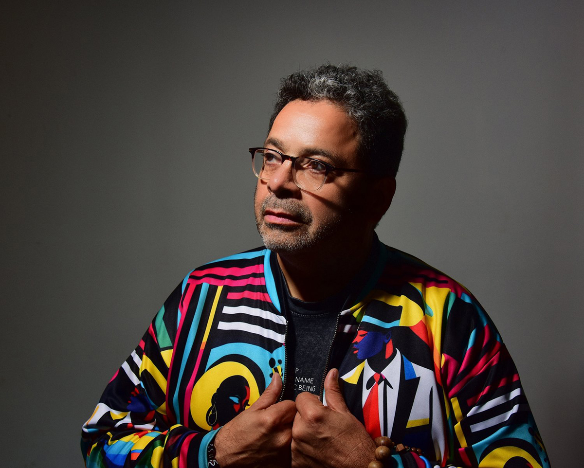 A man with short curly hair, beard, and glasses looks thoughtfully to the side. He wears a colorful patterned jacket over a dark shirt and stands against a plain gray background.