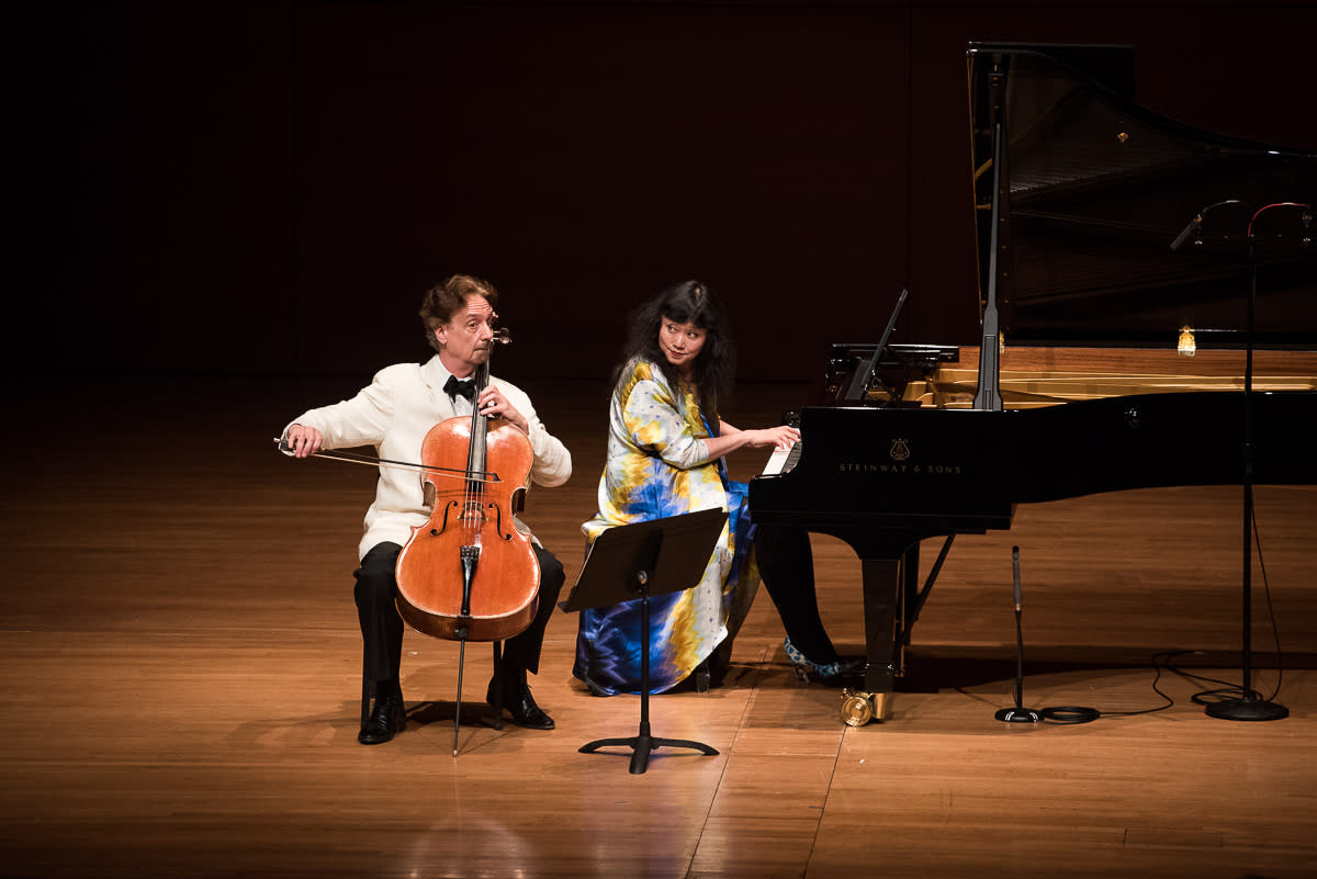 A man plays the cello while a woman in a blue and yellow dress plays a grand piano on a wooden stage, both focused on their performance, with sheet music on a stand between them.