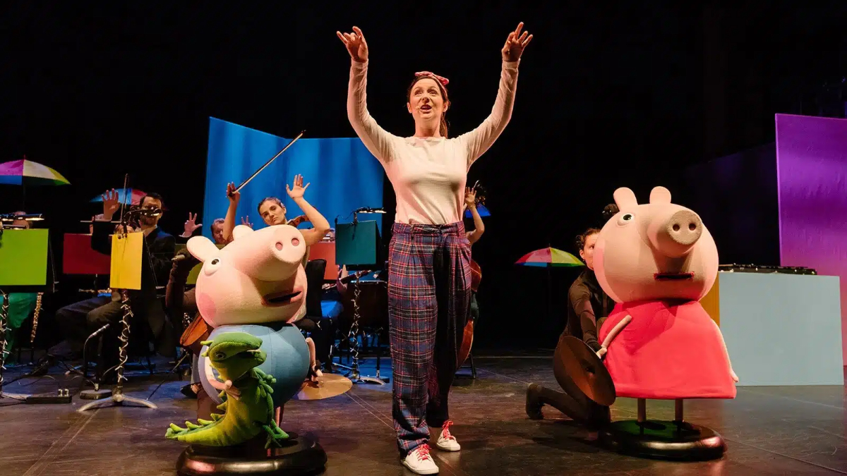 A performer in plaid pants stands on stage with arms raised, flanked by large Peppa Pig and George Pig costumes. Musicians play instruments in the background amid colorful set pieces.