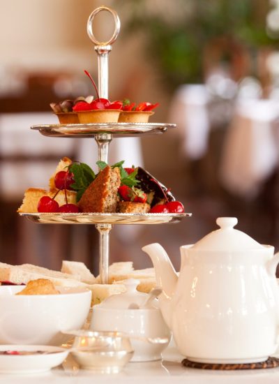 Elegant three-tiered high tea stand with pastries, tea sandwiches, cherries, and a white teapot set in a refined dining room setting.