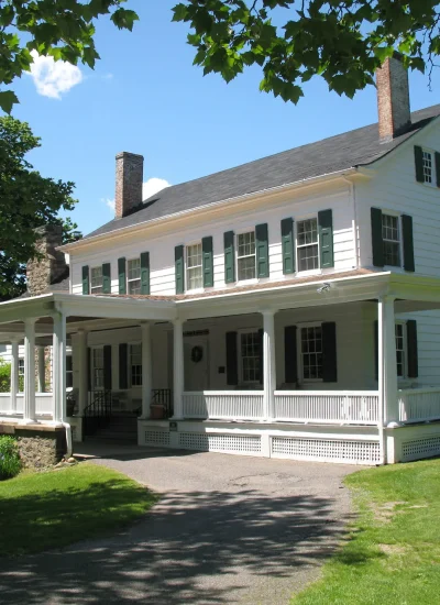 The historic white Sands-Willets House in Port Washington with green shutters, wraparound porch, chimneys, and surrounding trees on a sunny day.