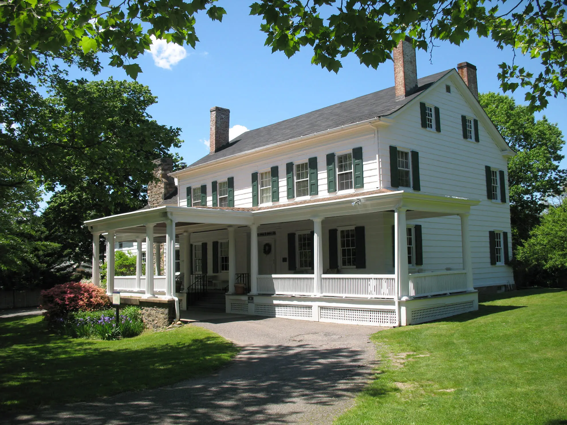 The historic white Sands-Willets House in Port Washington with green shutters, wraparound porch, chimneys, and surrounding trees on a sunny day.