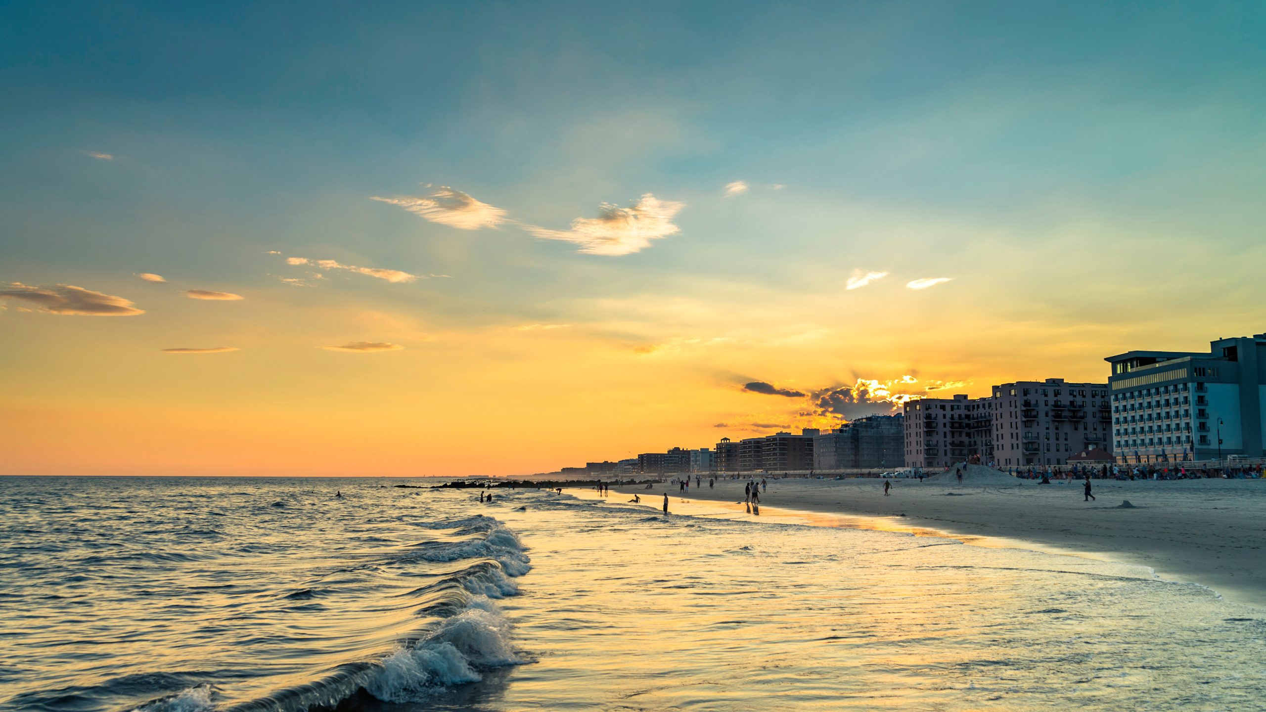 Golden sunset over a Long Island beach, with gentle waves rolling onto the shore and people walking along the waterfront near beachfront hotels and residences.