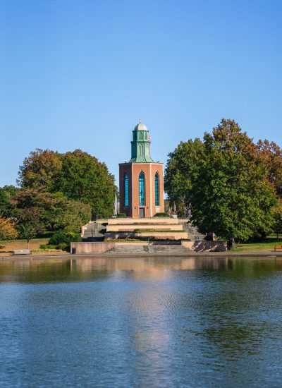 Eisenhower Park's iconic Harry Chapin Lakeside Theatre tower rises above a reflecting pond, framed by mature trees under a clear blue sky in Nassau County, New York.
