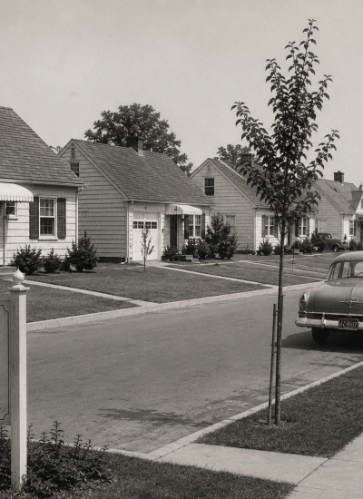 Vintage black-and-white photograph of a quiet Levittown, New York neighborhood in the late 1940s, featuring rows of Cape Cod-style homes, classic automobiles, and a mother walking with her child along a tree-lined sidewalk.
